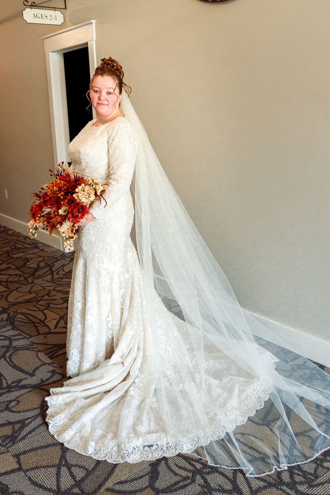 Bride in lace wedding gown with long train and veil holds autumn-colored bouquet.