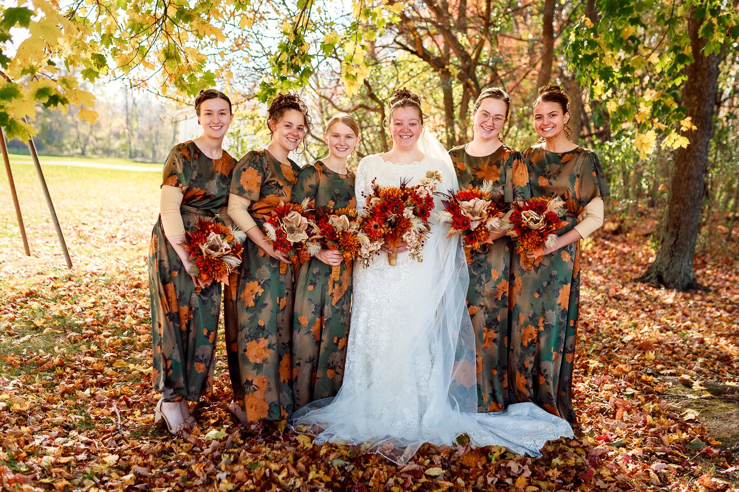 Wedding party poses outdoors with bridesmaids in floral dresses against autumn trees.