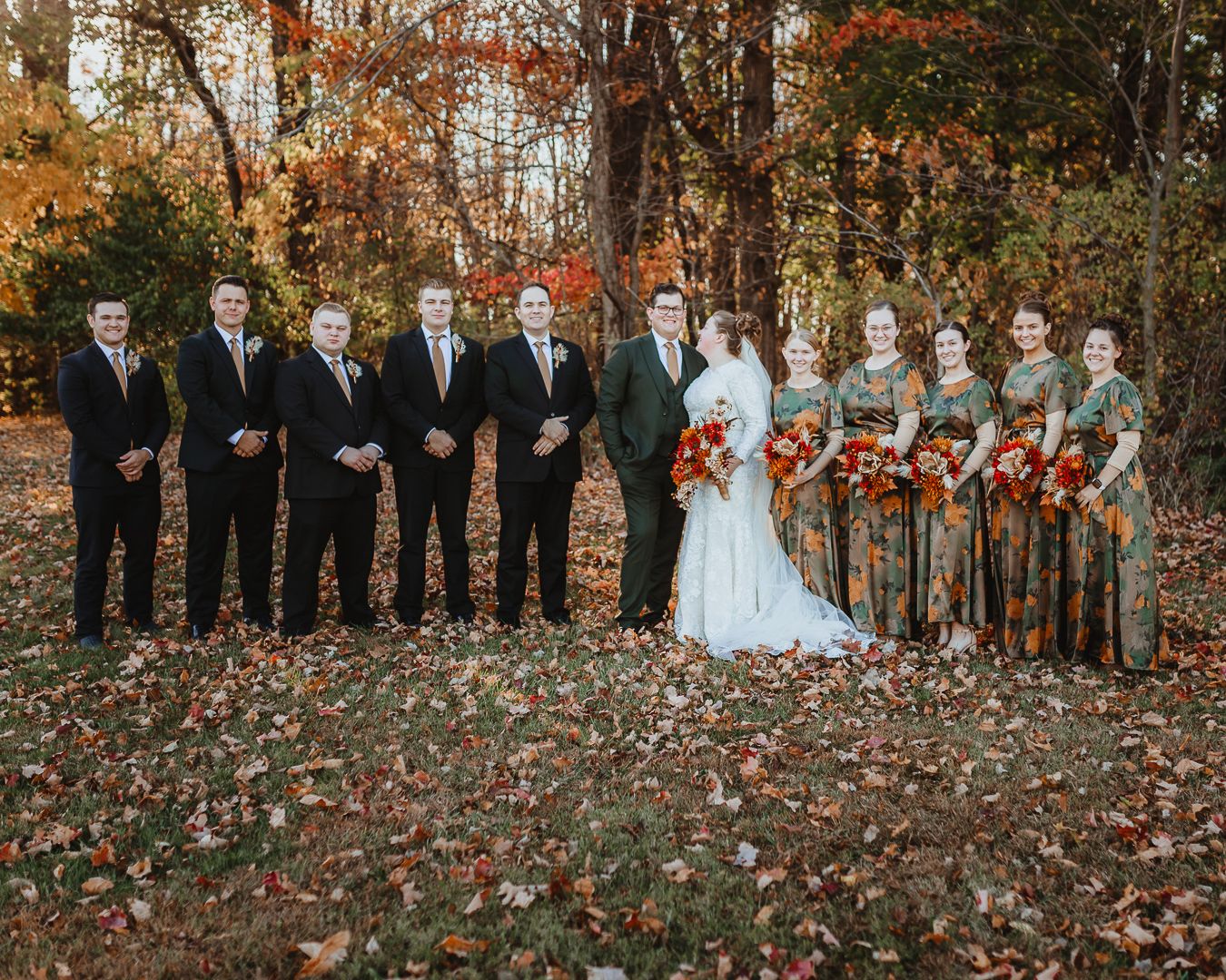 Large wedding party lined up for formal photos among fall foliage.