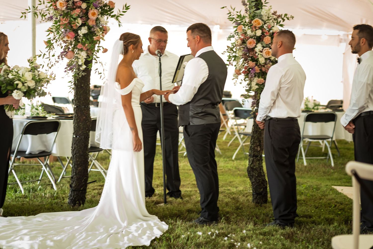 Outdoor wedding ceremony under a white tent with floral arrangements and chairs set up on grass.