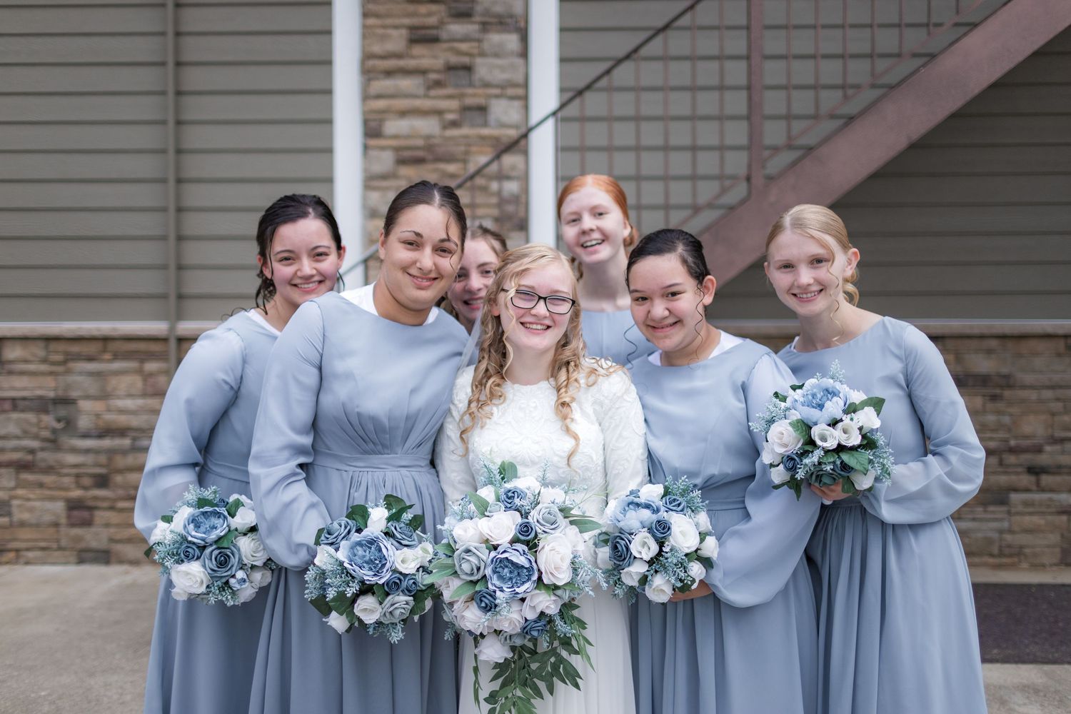 Bridesmaids in light blue dresses pose together with bouquets against a stone and grey building.