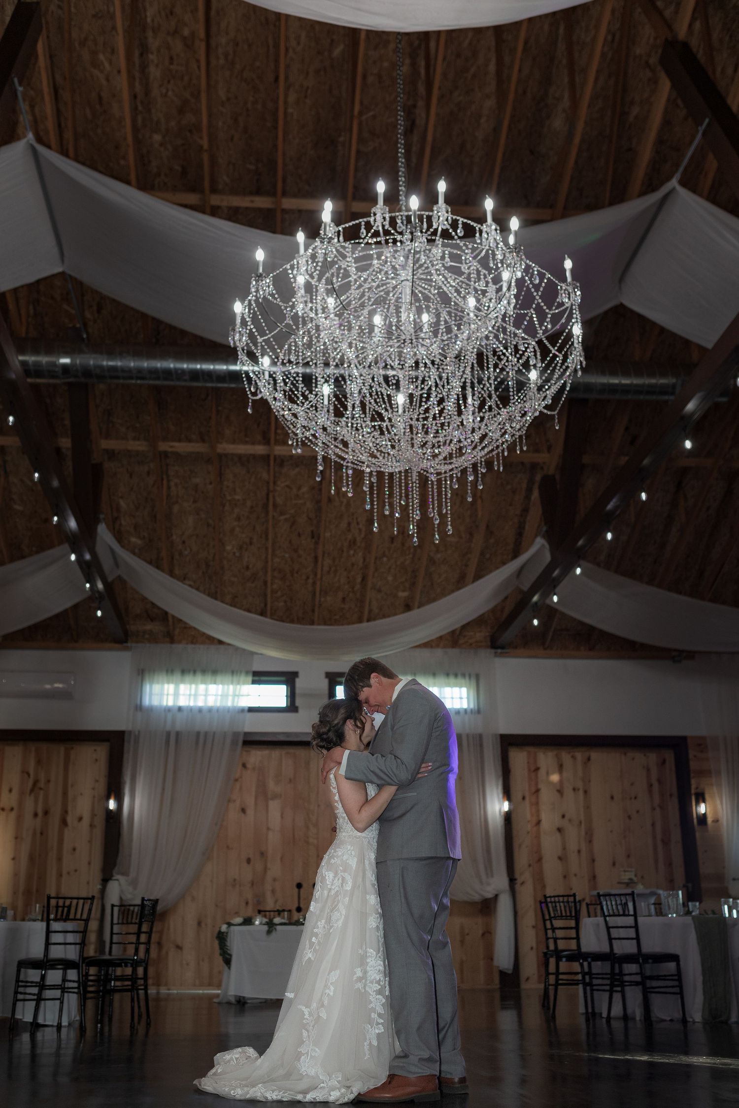 Elegant crystal chandeliers illuminate a rustic barn wedding venue with wooden beams.