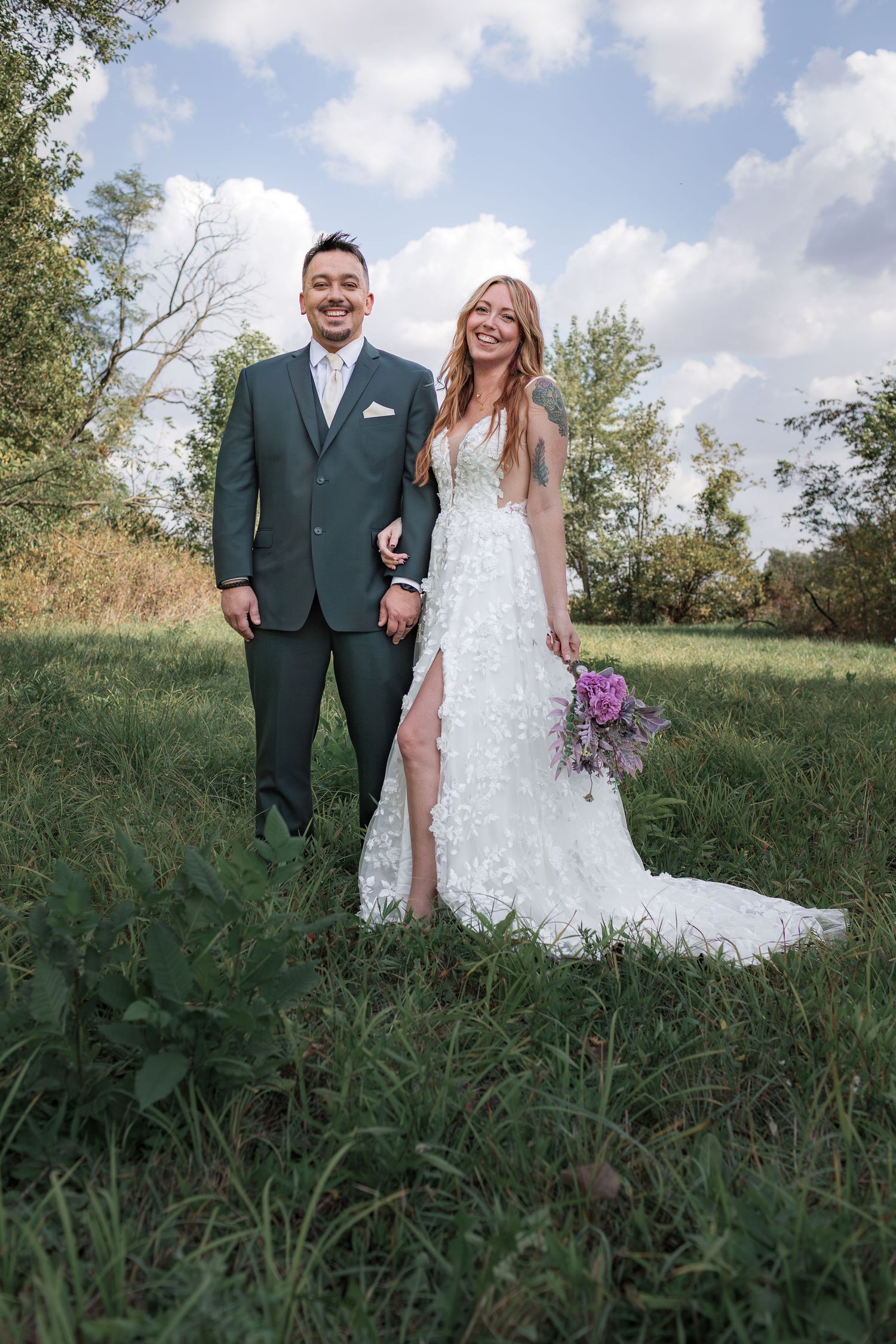 A couple poses in a grassy field on their wedding day, with the bride wearing a white lace gown and holding purple flowers.