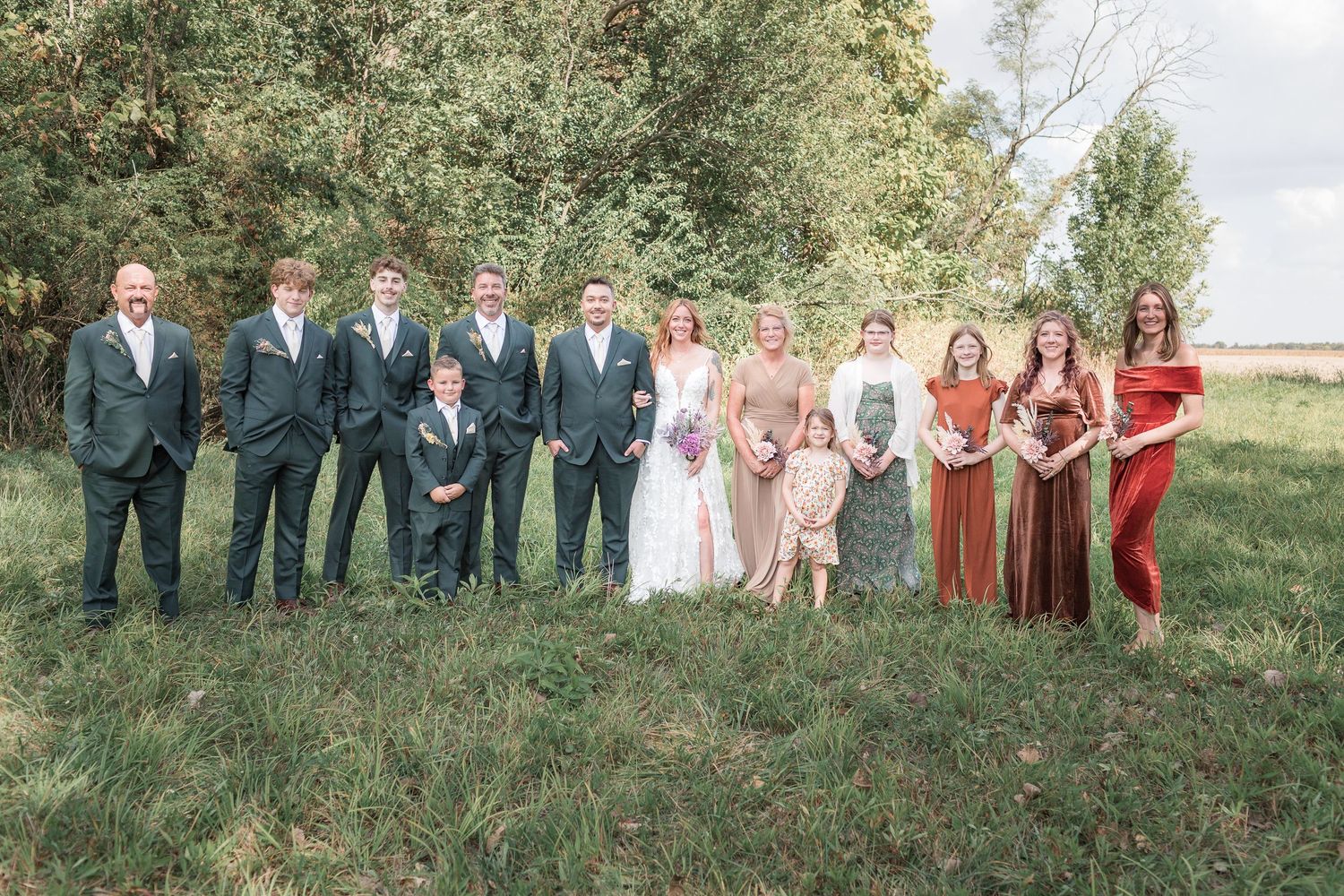 Large wedding party in coordinated attire poses together on a grassy hillside.