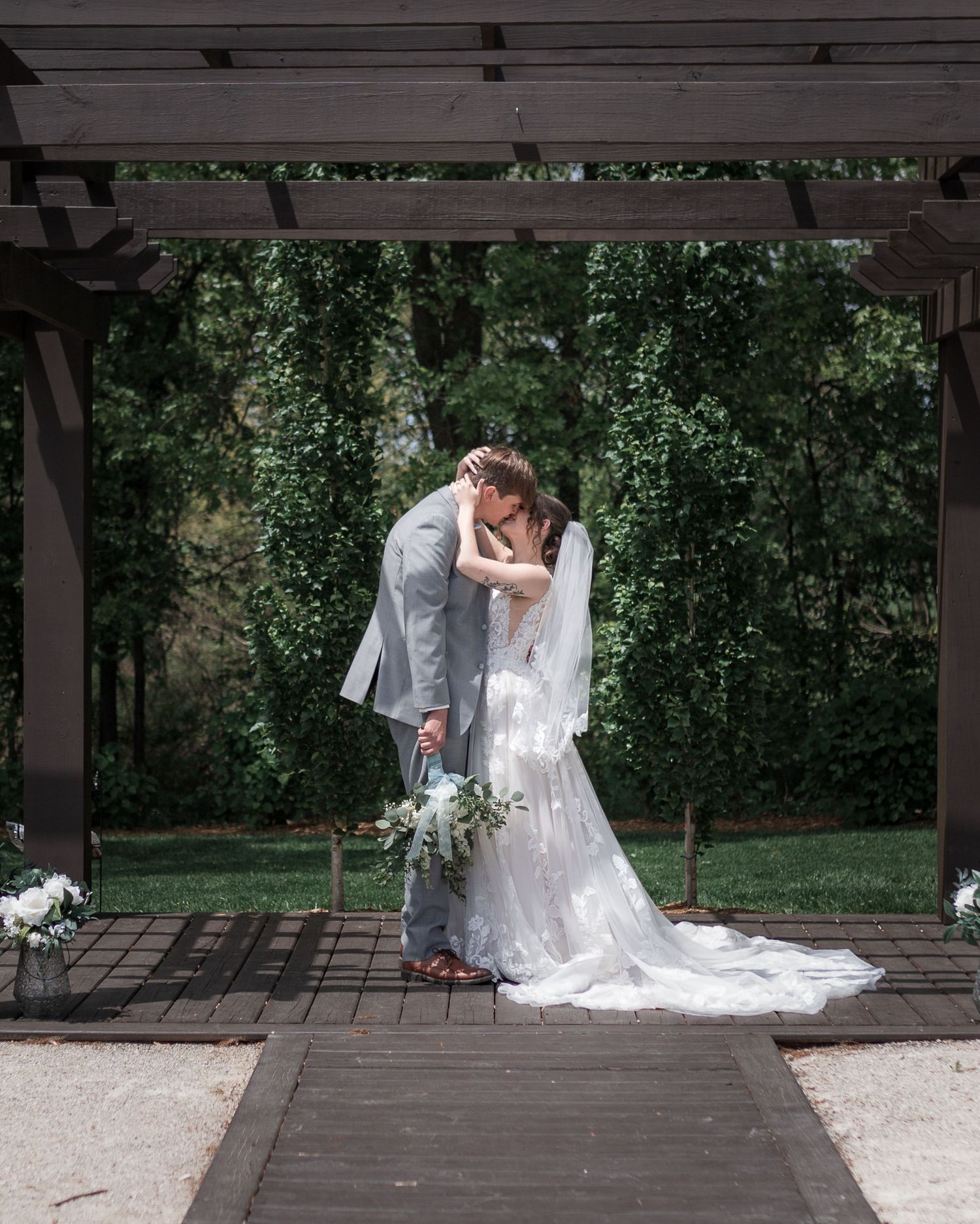 Romantic wedding couple shares intimate moment under wooden pergola in garden setting.