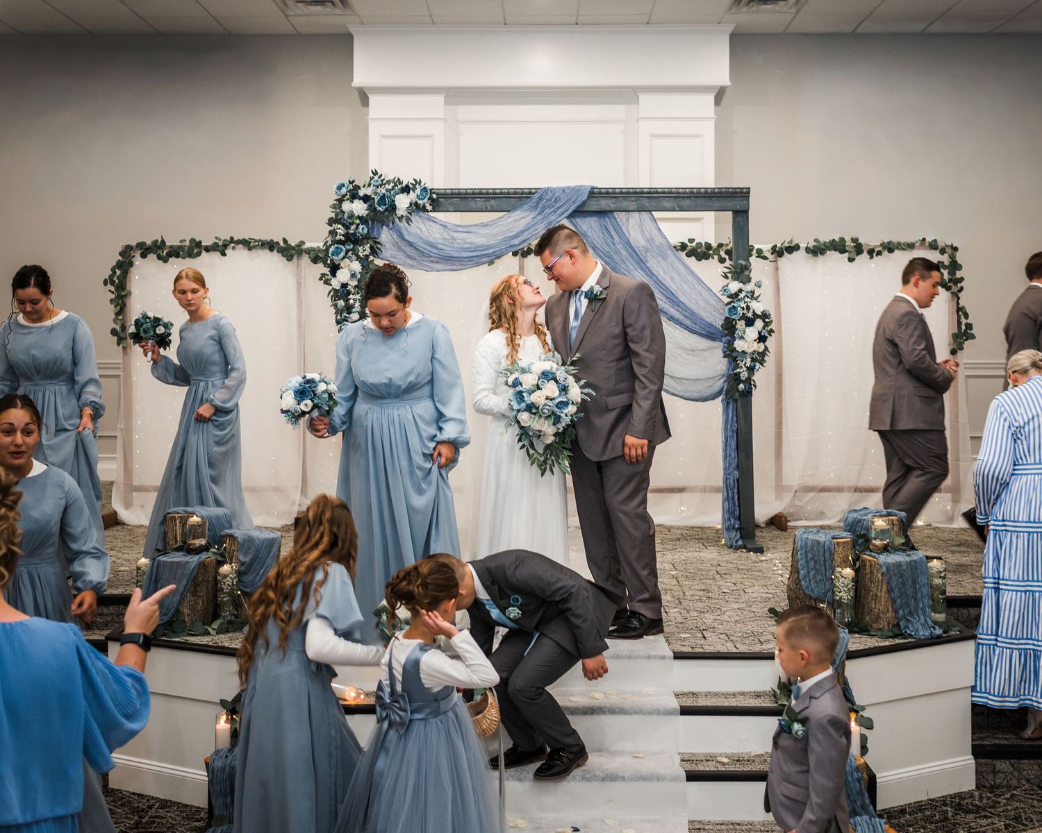 Wedding party in blue and grey attire poses and celebrates in an indoor venue with floral decorations.