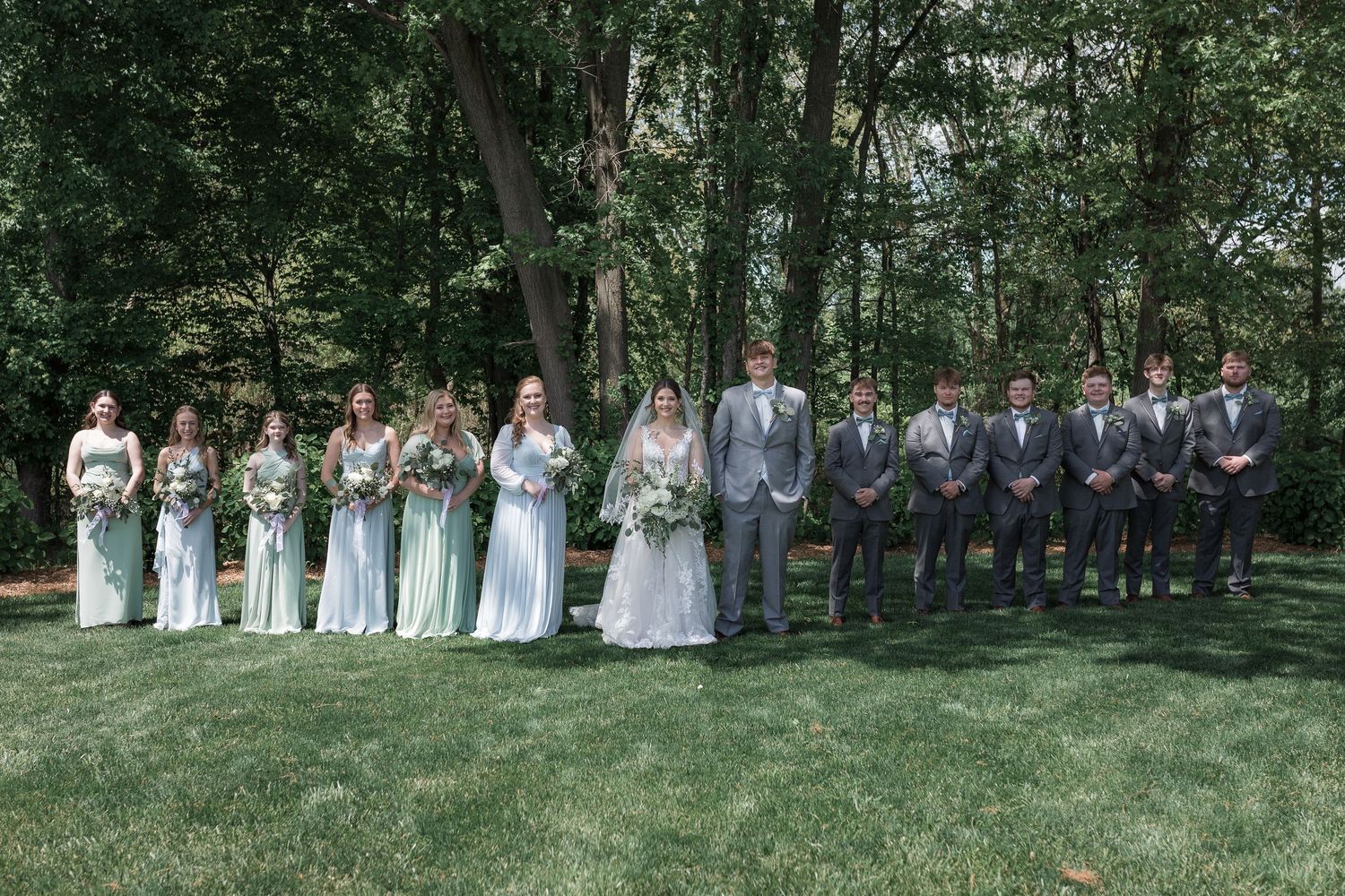 Wedding party poses together on grassy lawn with bridesmaids in light blue dresses and groomsmen in gray suits.