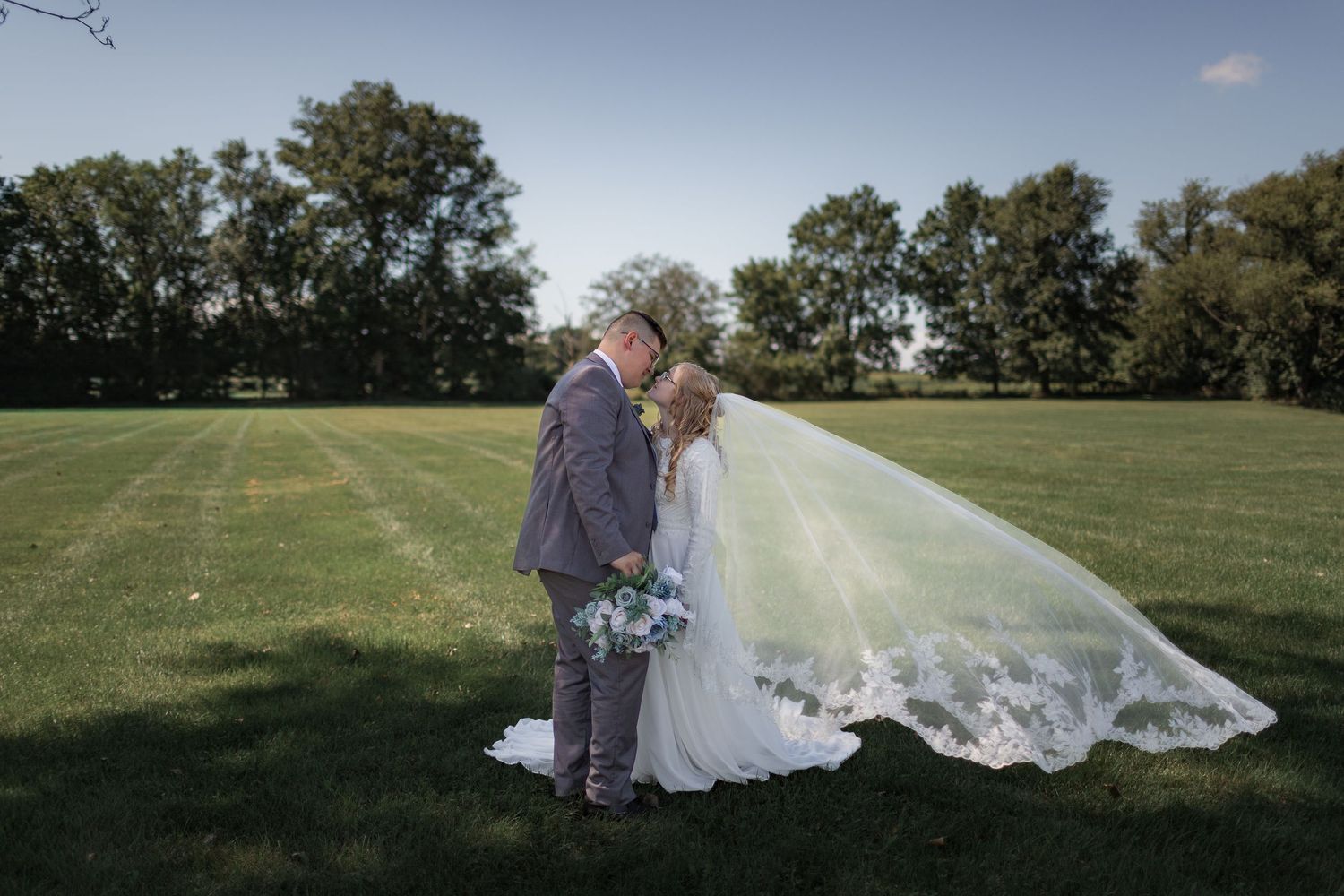 Bride and groom embracing on a grassy field with trees in the background.