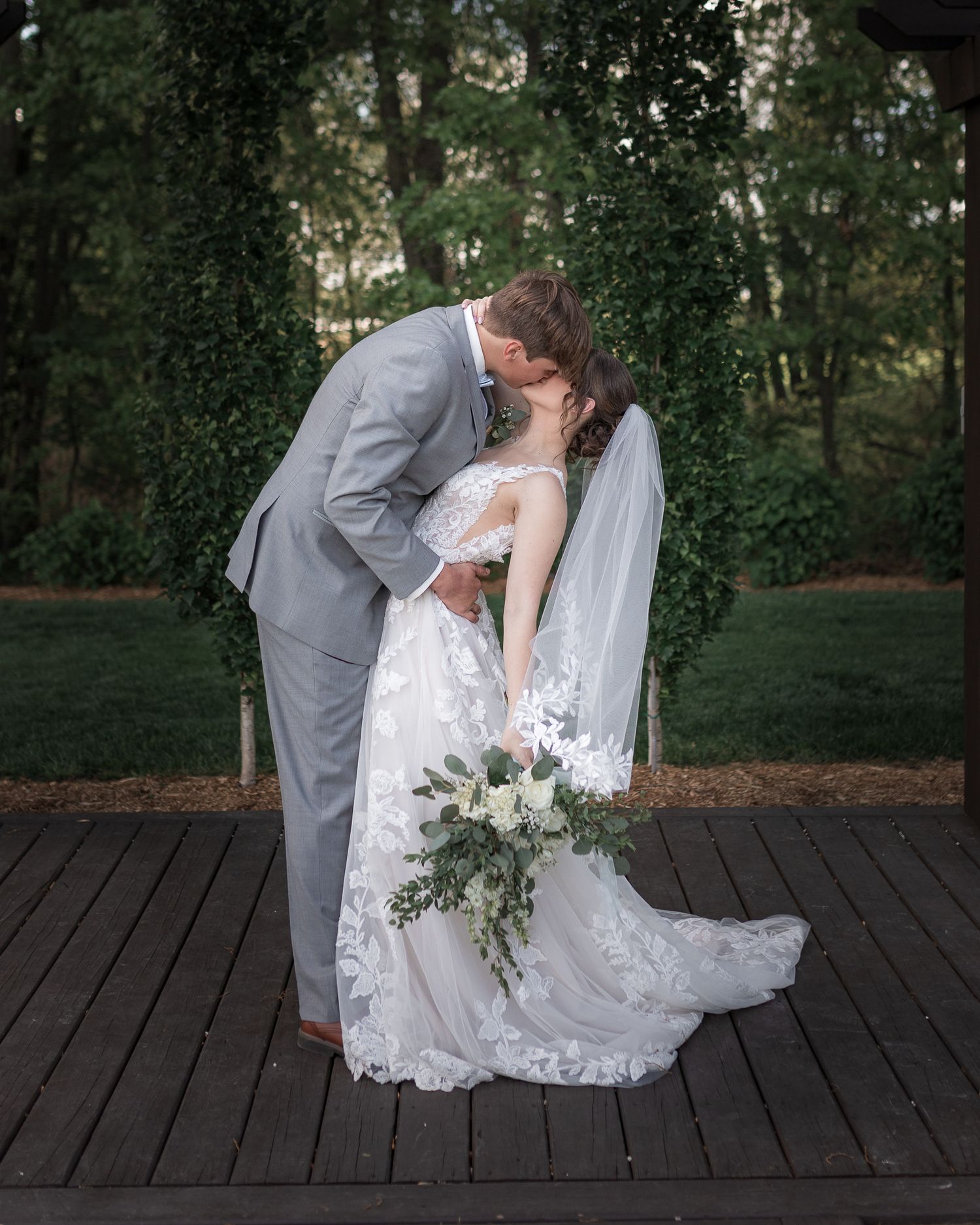 Newlyweds share intimate moments on wooden deck surrounded by natural greenery.