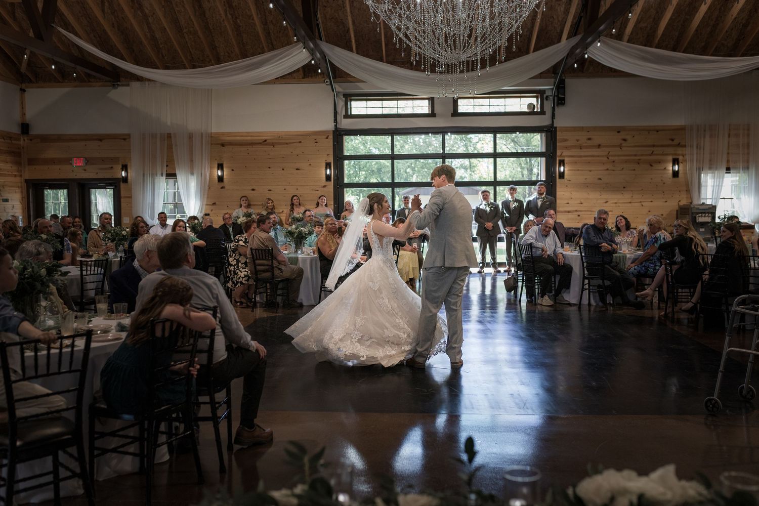 Wedding reception venue with rustic wooden beams and large windows filled with celebrating guests.