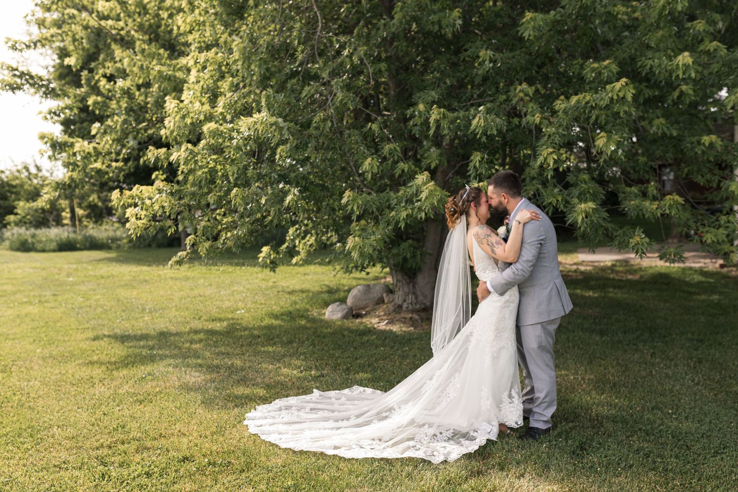 A bride and groom share a romantic kiss on a lush green lawn with trees in the background on their wedding day.