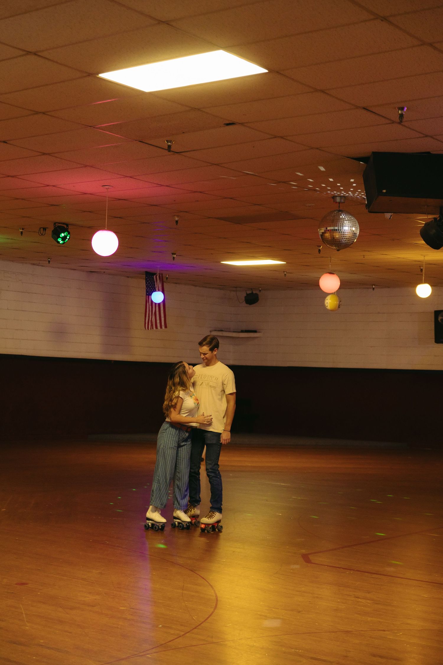 A couple roller skating together in an indoor rink with disco lights and decorative globe lights hanging from the ceiling.