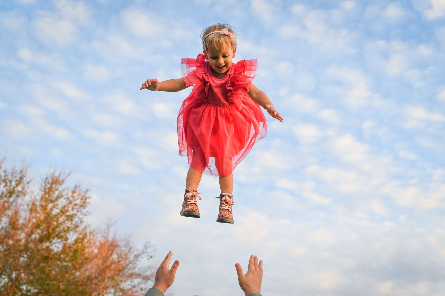 A small figure in a flowing red dress and sandals appears suspended mid-air against a blue sky with autumn trees below.