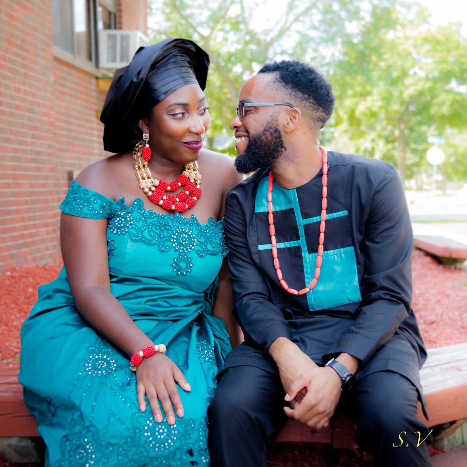 A couple in coordinating turquoise and black outfits with beaded necklaces sit together on a bench near brick building.