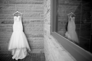 White wedding dress hangs against stone wall with reflection in window.