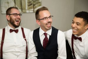 Three groomsmen in bowties and suspenders share a candid laughing moment.