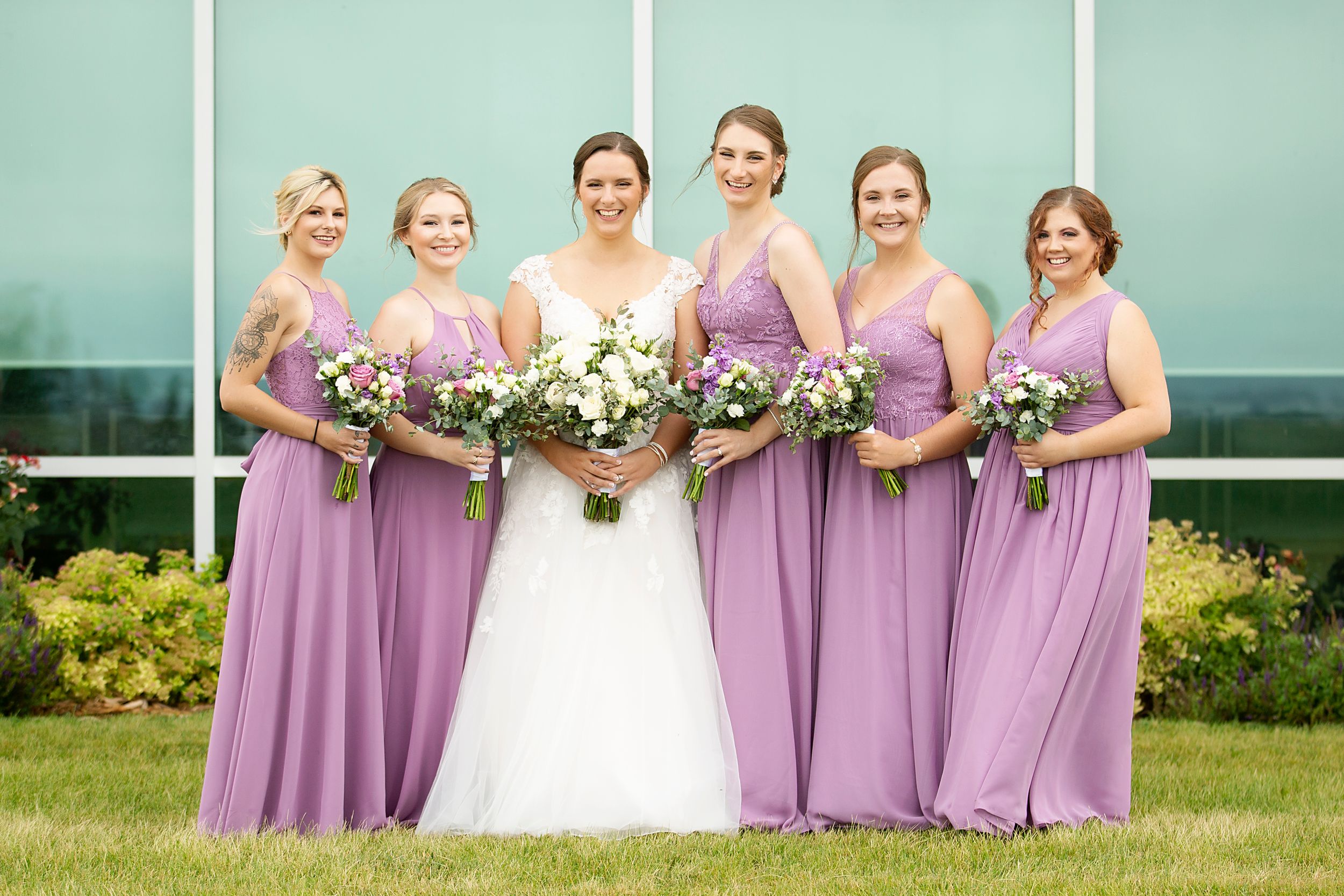 Bridal party in lavender dresses poses with bride in white gown against turquoise wall.