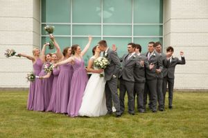 Wedding party in purple bridesmaid dresses and gray suits pose playfully against turquoise wall.