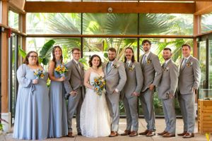 Wedding party in gray suits and lavender dresses poses under tropical palm covering.