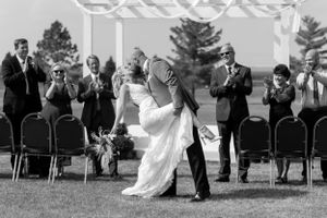 Black and white photo of a wedding ceremony kiss under a white pergola with guests applauding.