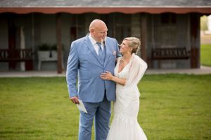 A joyful wedding couple in light blue suit and white lace dress share a candid moment on a grassy lawn.