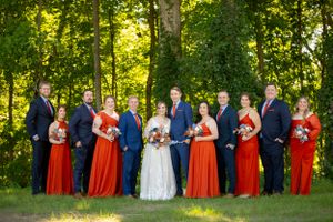 Wedding party in navy suits and vibrant red dresses pose together on a sunny lawn with trees.