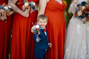 A young ring bearer in a navy suit poses with bouquets among red bridesmaid dresses.