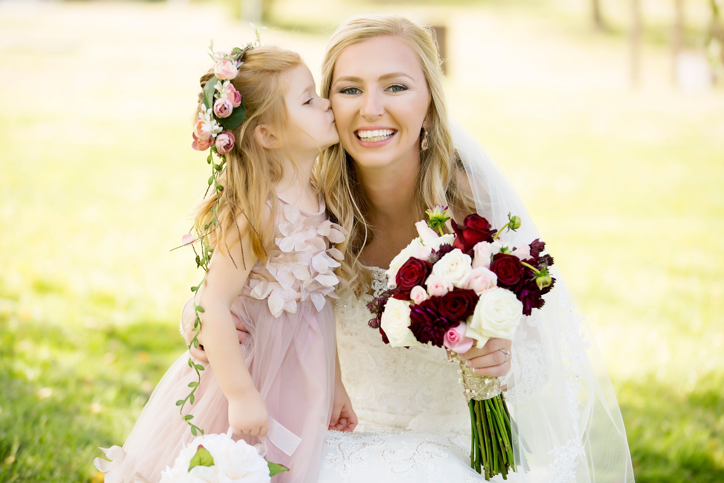 Candid outdoor wedding portrait with red and white bouquet in sunlit garden setting.