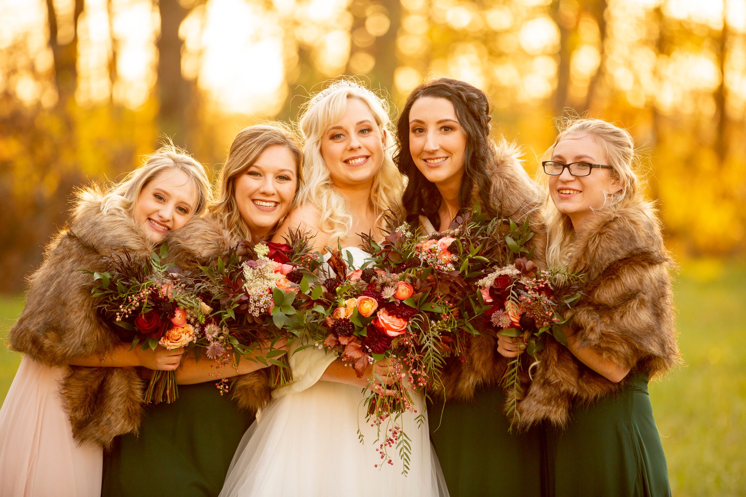 Bridal party wearing fur stoles poses with bouquets in golden autumn light.