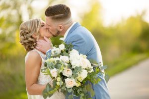 A romantic kiss between newlyweds holding a lush white and green bouquet outdoors.