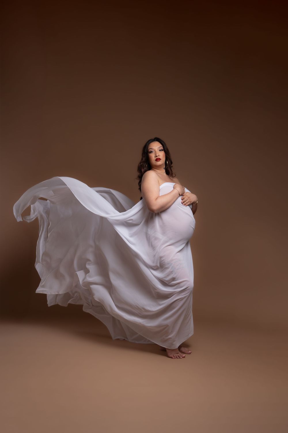 Photo de maternité élégante avec une robe en tissu blanc fluide sur fond marron dans un portrait artistique en studio.