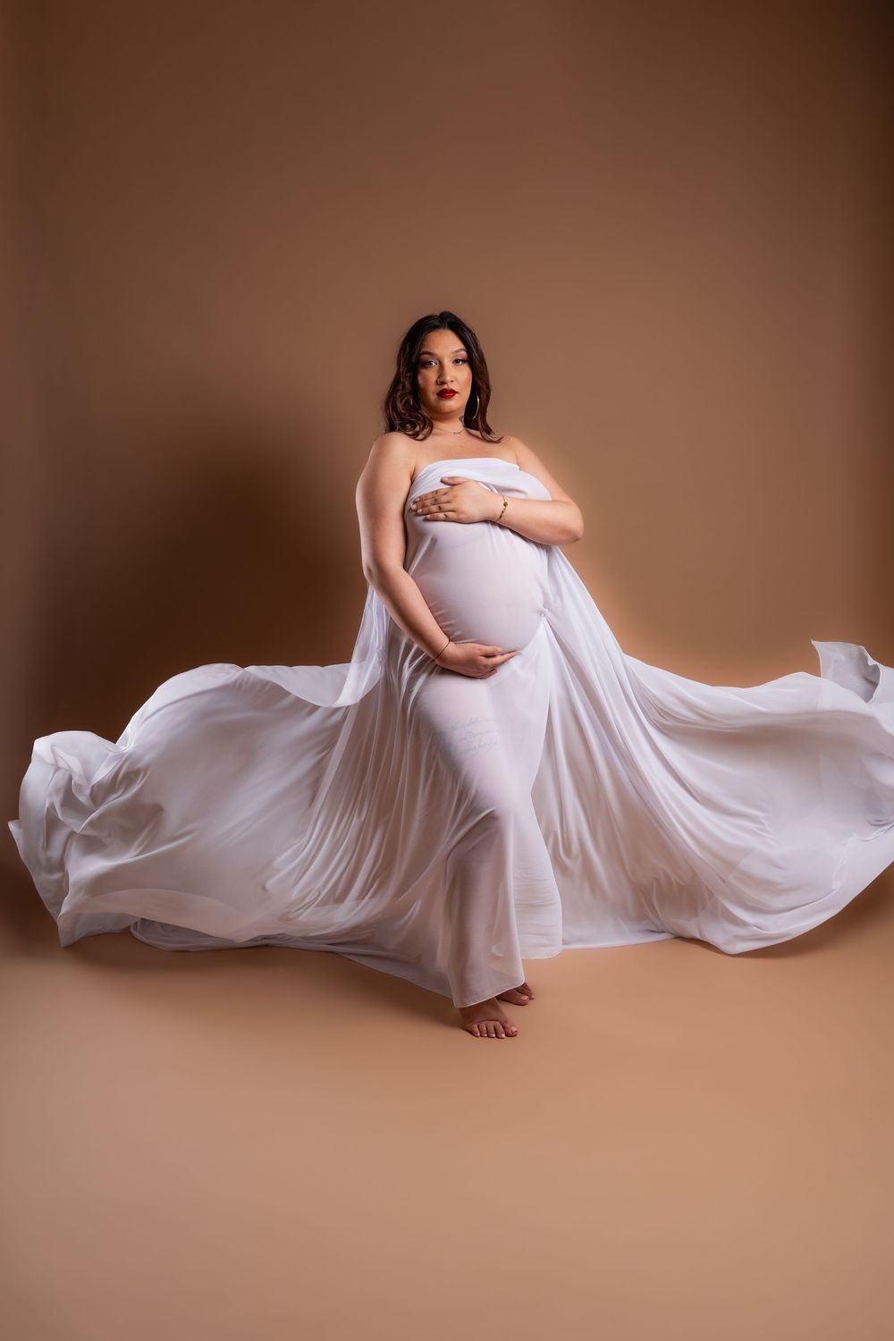 Une future maman en robe blanche fluide pose élégamment sur fond marron lors d'une séance photo de maternité.