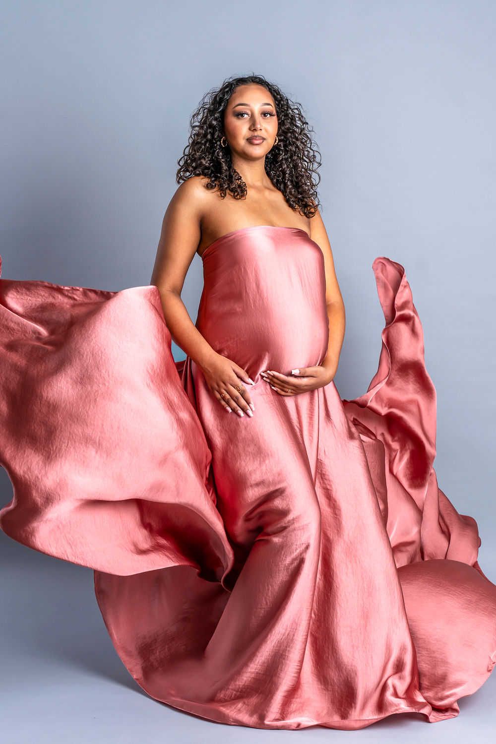 Une belle robe de maternité en soie rose fluide ondule de façon spectaculaire pendant une séance photo de grossesse sur fond gris clair.