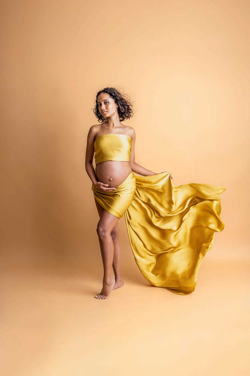Une séance photo de maternité présente une robe en soie jaune fluide sur un fond beige chaud, créant une silhouette élégante.