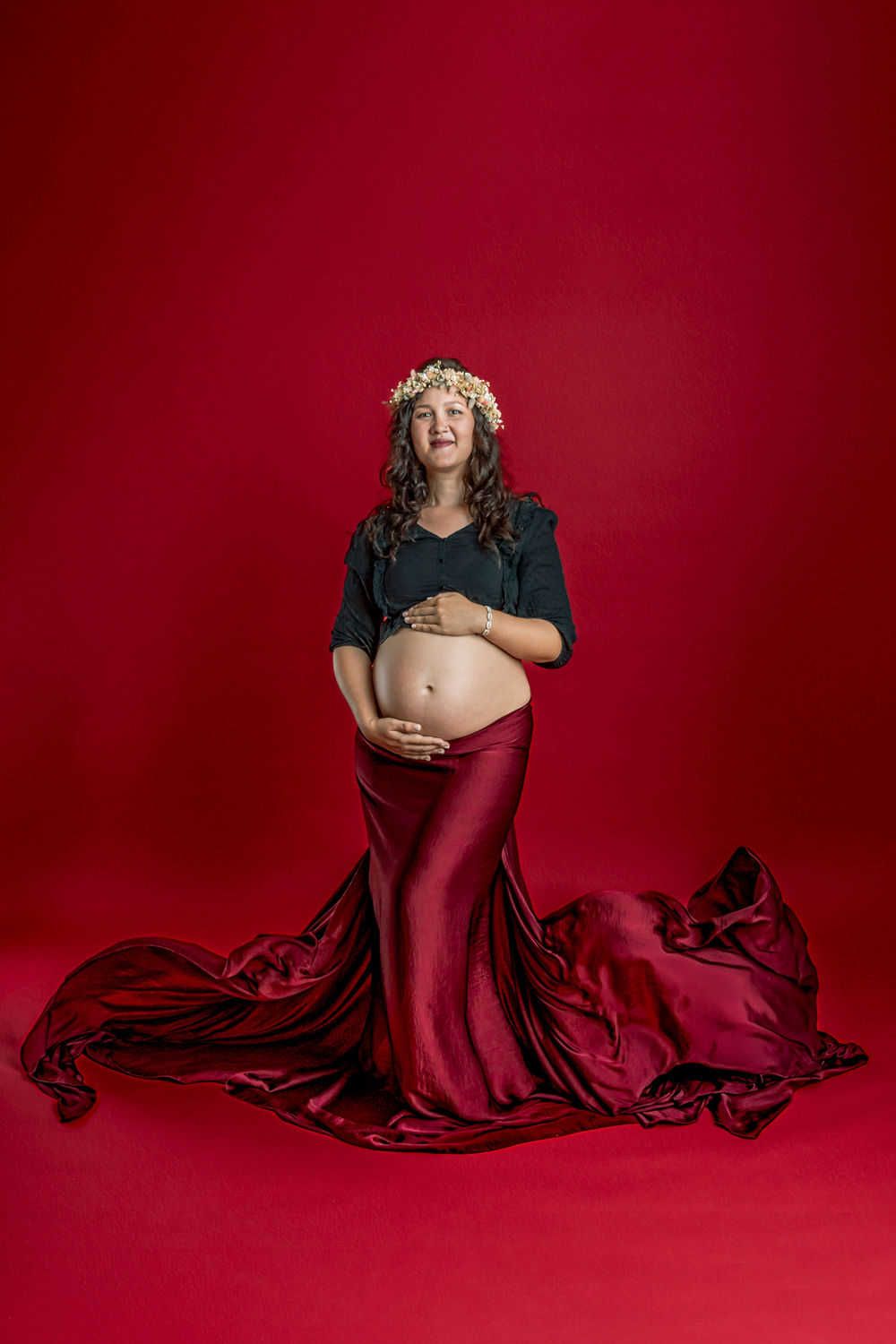 Portrait de maternité sur fond rouge avec robe fluide en soie bordeaux et couronne florale sur fond cramoisi foncé