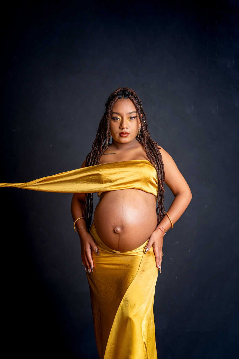 Une femme enceinte portant une robe jaune fluide pose sur un fond sombre lors d'une séance photo de maternité artistique.