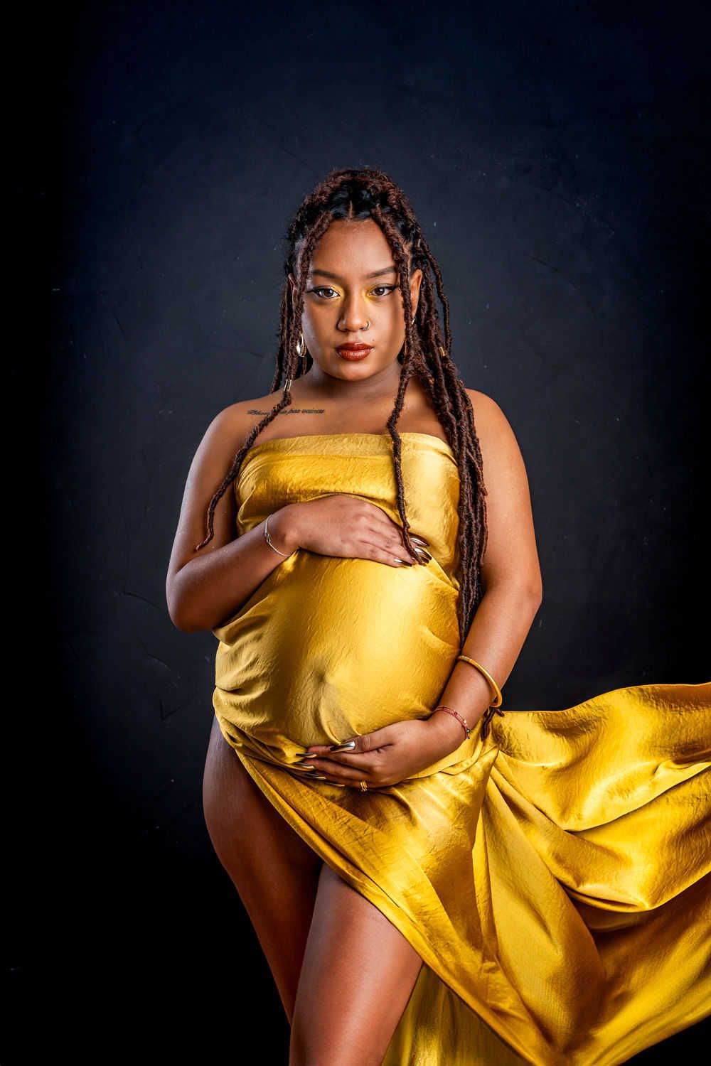 Élégante photo de maternité mettant en vedette une robe fluide en soie dorée sur fond sombre dans un éclairage de studio dramatique.
