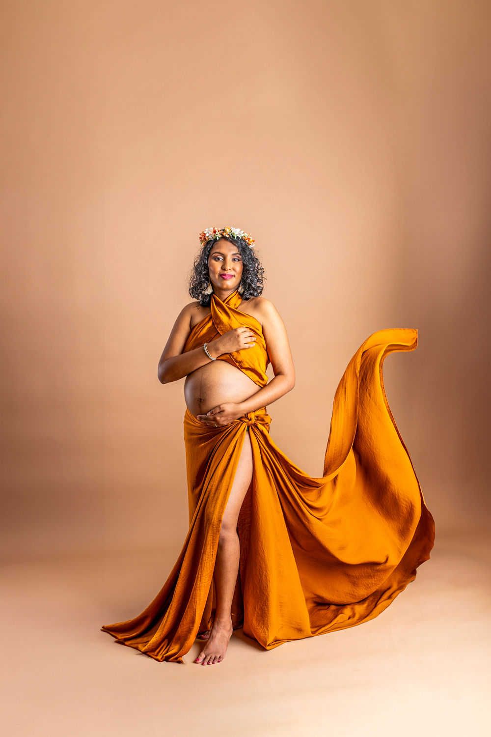 Une femme enceinte vêtue d'une robe orange fluide pose gracieusement sur fond beige lors d'une séance photo de maternité.