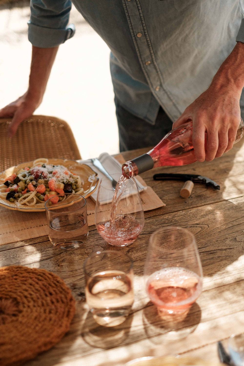 Hands pouring rose wine into glasses on a wooden table with food and woven placemats.