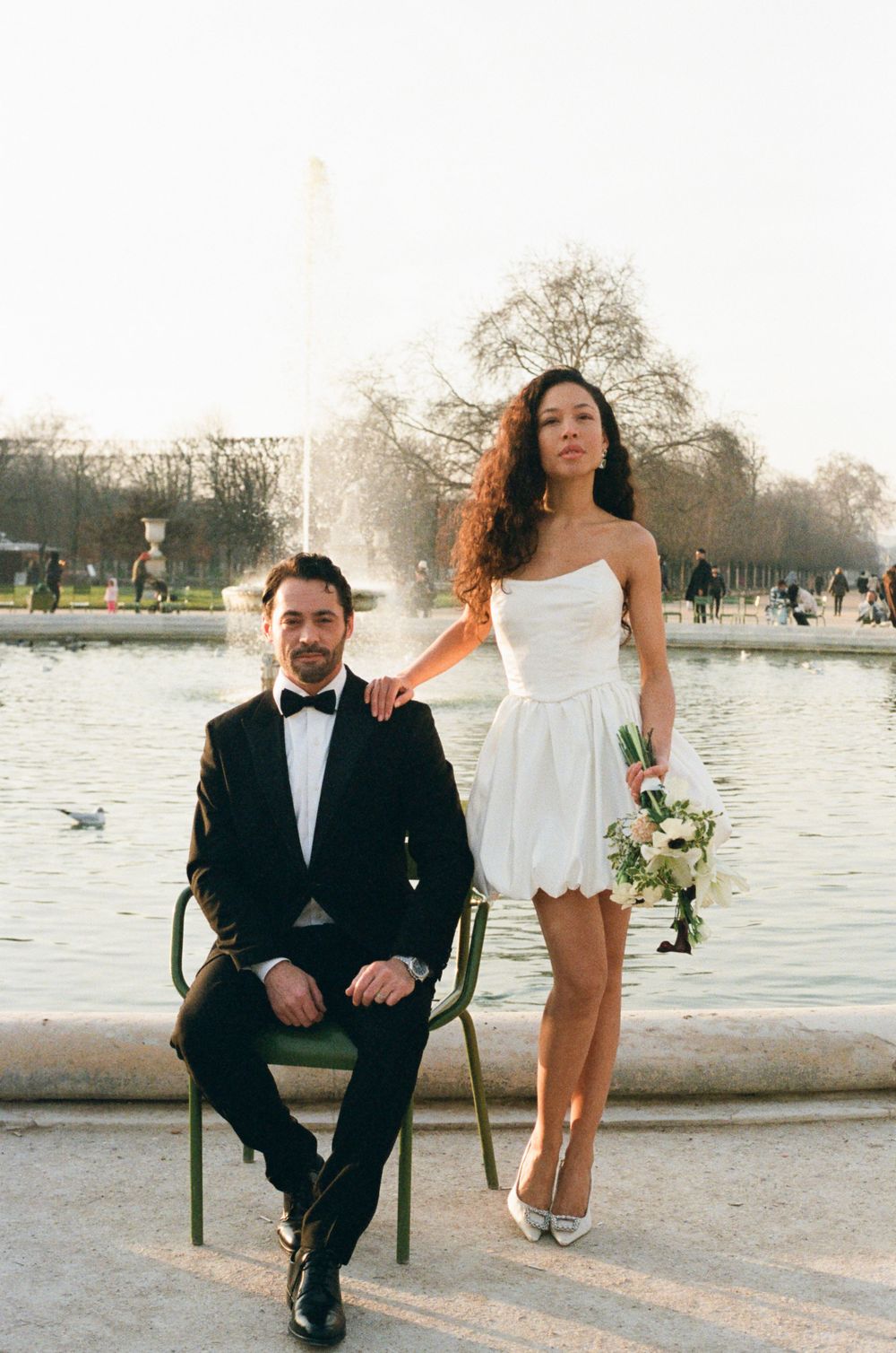 A couple poses near a pond in Paris, with the bride in a short white dress and groom in black tuxedo.