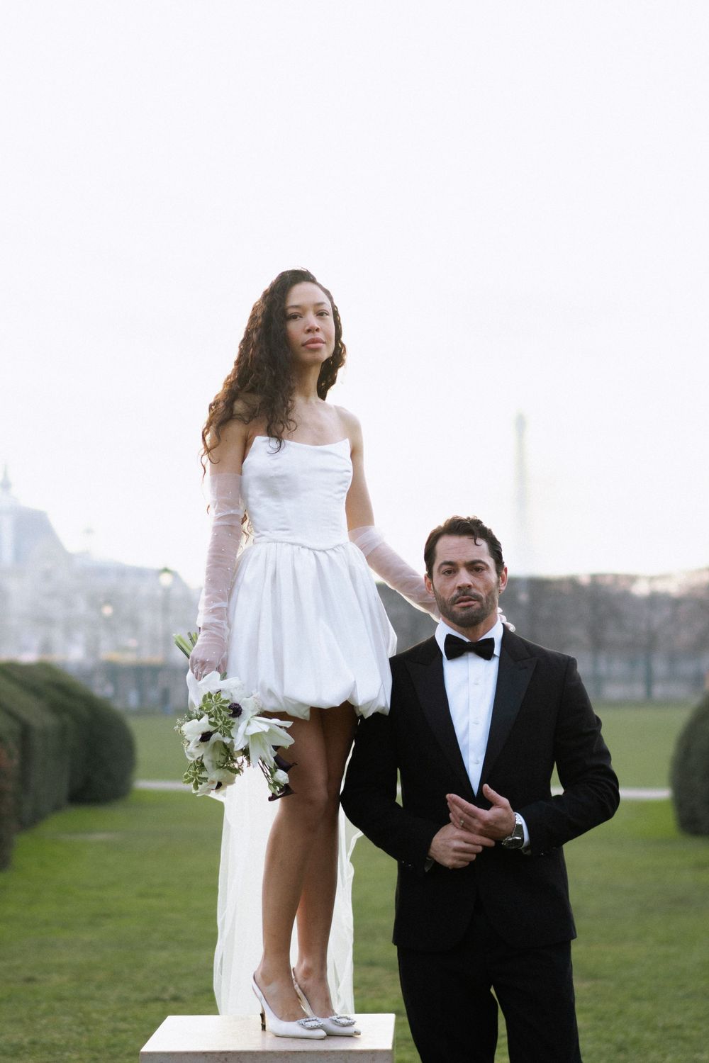 A couple in wedding attire pose outdoors on a green lawn, with the bride standing on a platform in a short white dress.