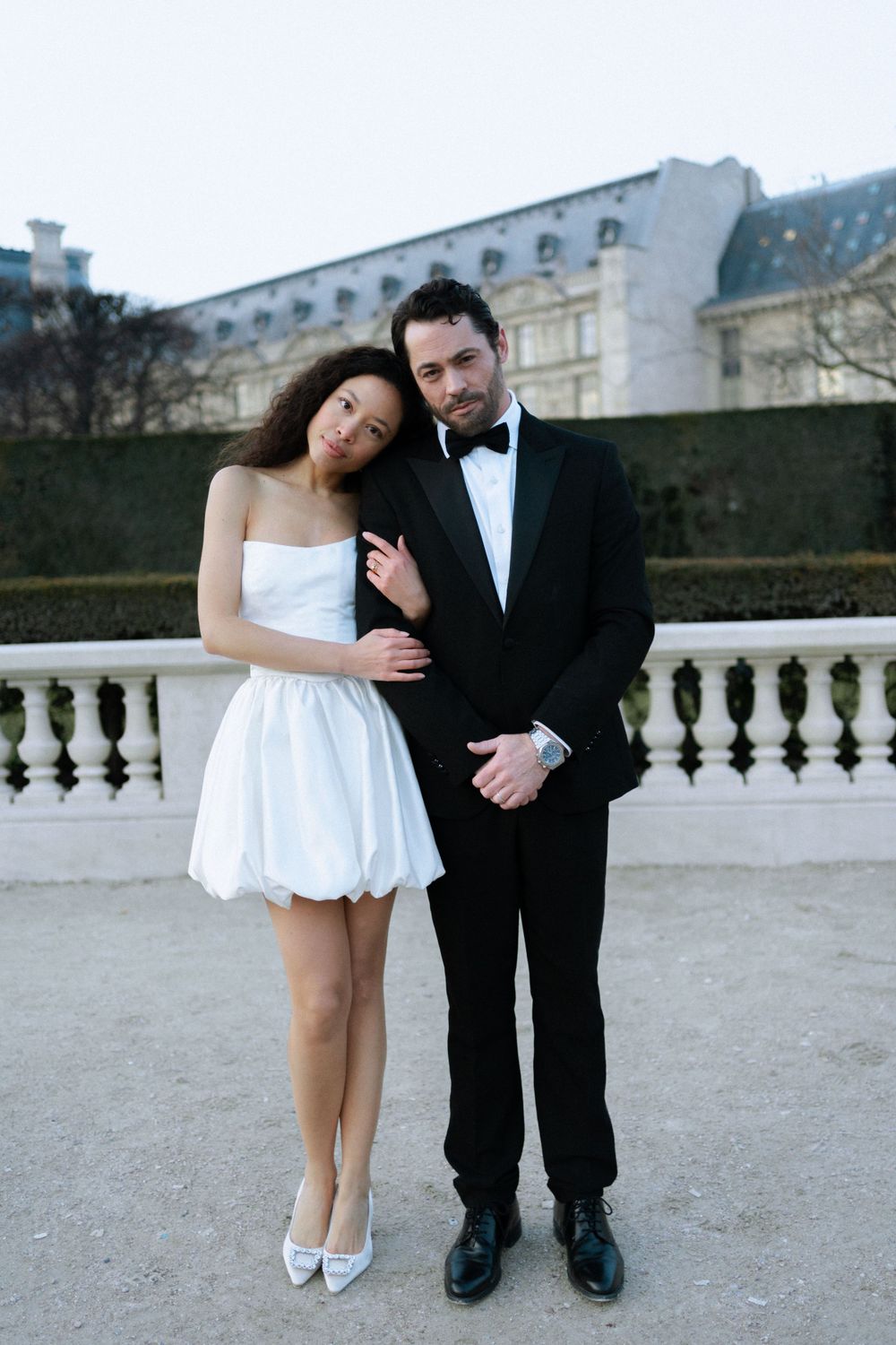 A couple poses in formal attire against an elegant balustrade with a historic building facade in the background.