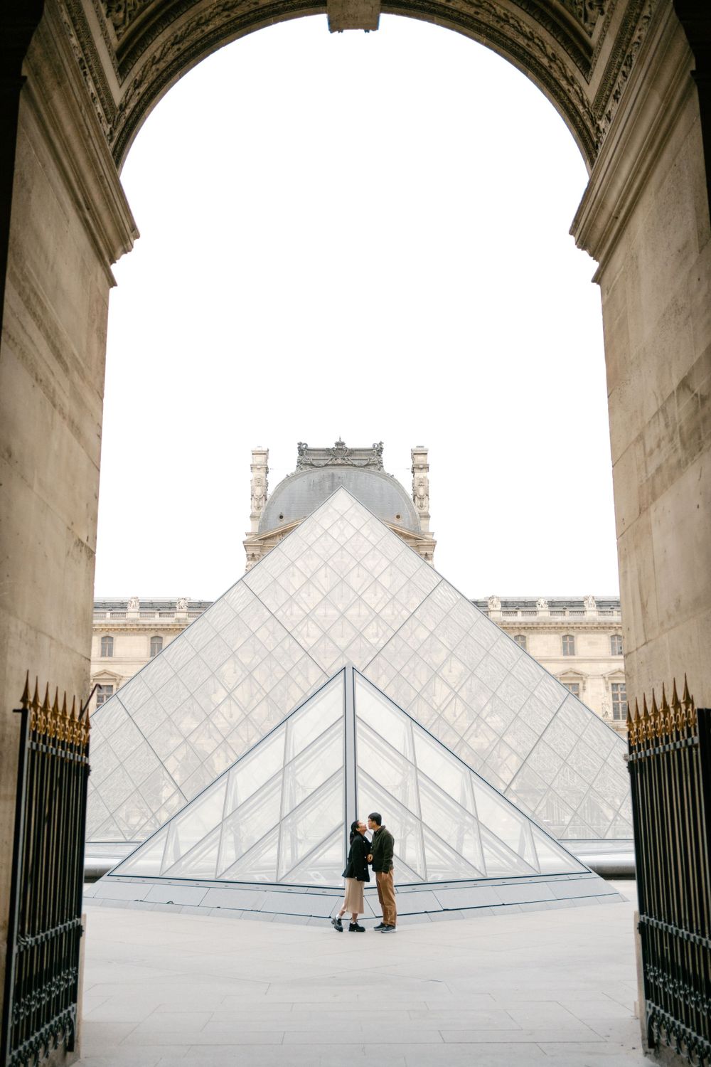 A romantic silhouette under an archway at the Louvre Museum with the iconic glass pyramid in the background.