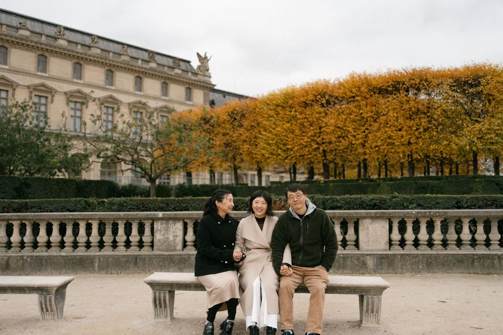 Three people pose on stone balcony with golden autumn trees and historic building facade in Paris.