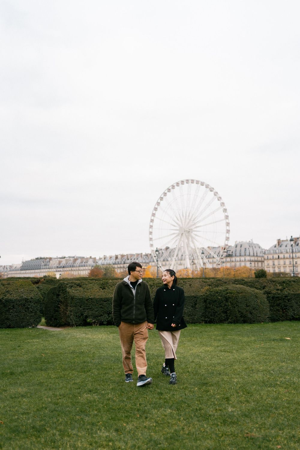 Two people walking on grassy lawn with Parisian Ferris wheel and classic buildings visible in background.