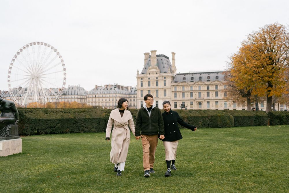 Three friends walking on grass near Louvre Museum with ferris wheel visible in autumn setting.