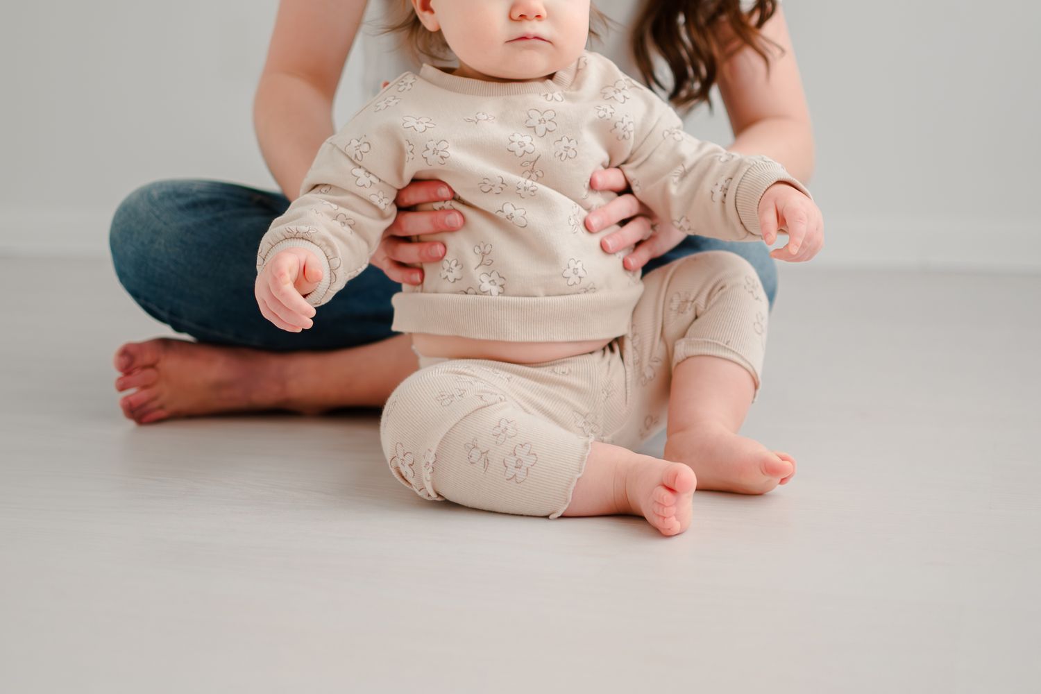 A baby wearing a cream-colored sweater sits on a light gray floor while held by someone in jeans.