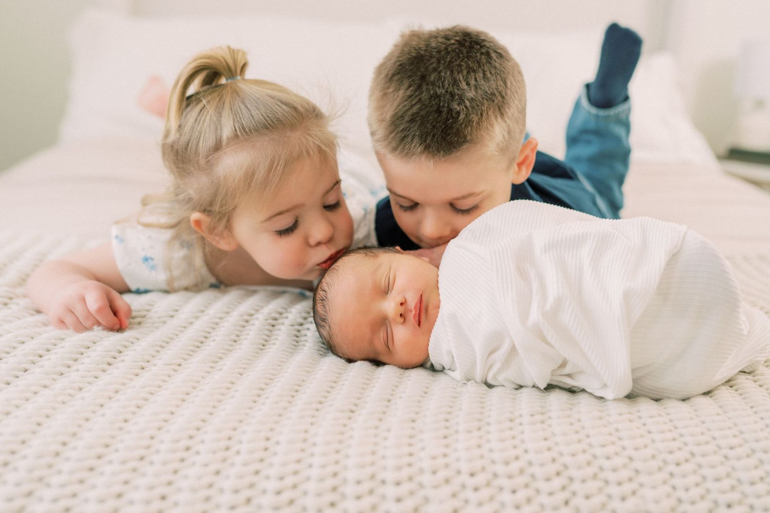 Family relaxes together on a white bed in a bright, sunlit bedroom with hardwood floors and white decor.