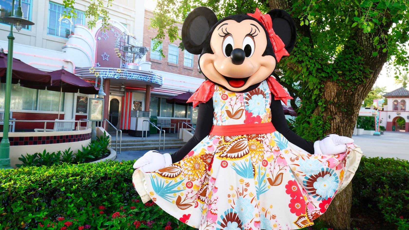Mickey Mouse character poses in colorful floral dress at Disney park with vintage-style buildings in background.