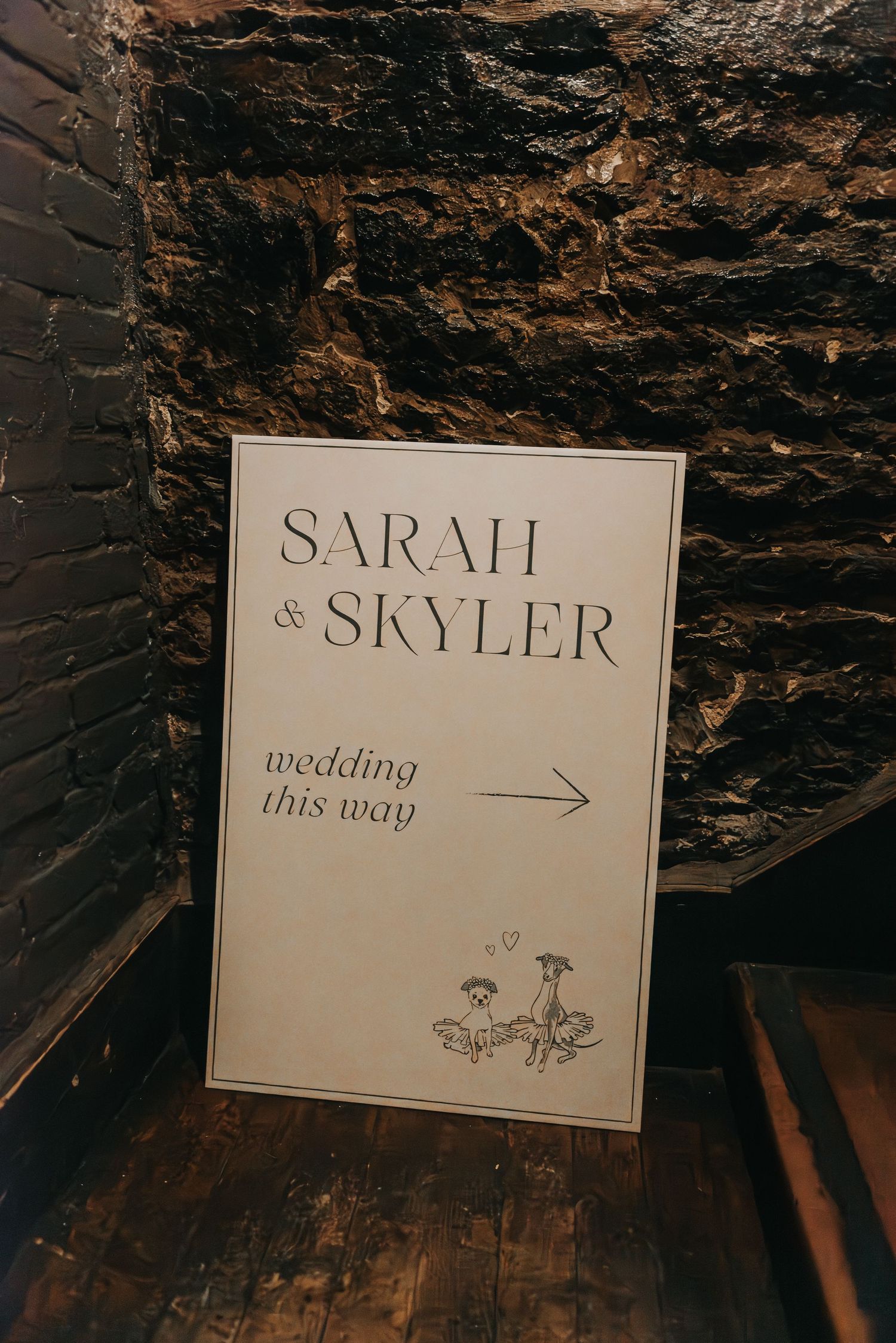 An elegant wedding directional sign on white paper with floral details mounted against a dark textured stone wall.